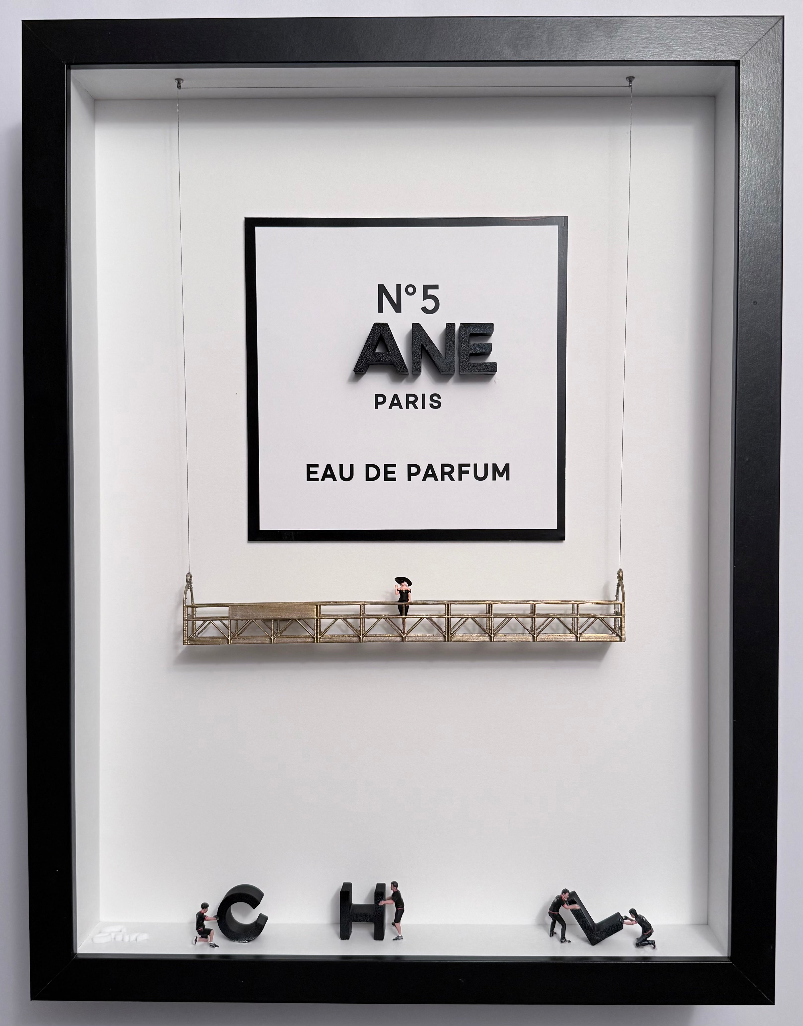 Shadow box artwork of a partially installed perfume logo CHANEL. Workers prepare to mount the missing letters while a woman in black outfit and hat stands on a gold platform. 