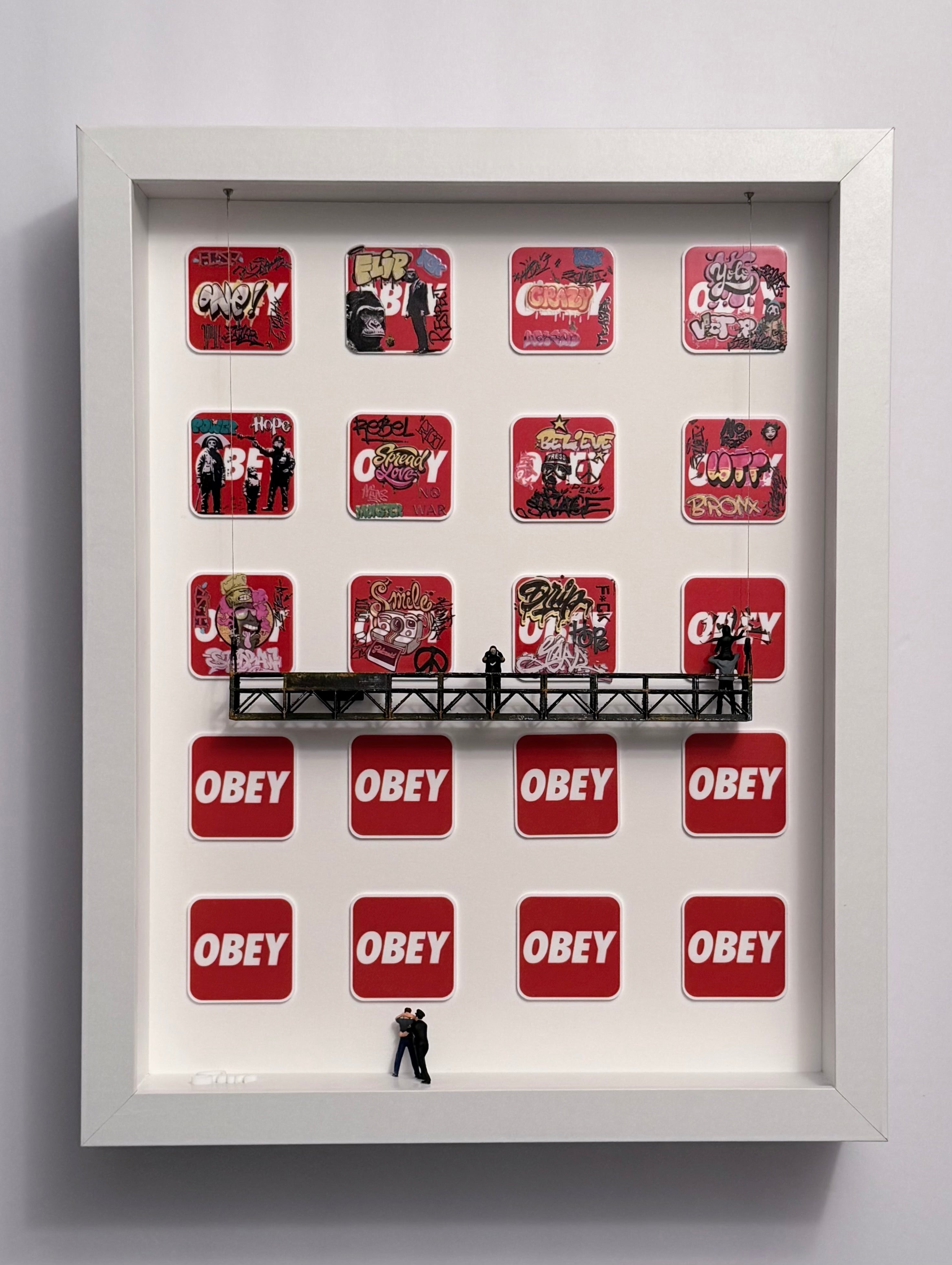 Rows of word obey form a backdrop to a rebellious scene where graffiti and defiance clash with control. A man raises his fingers in protest as another paints over authority is standing on a platform with two other miniature figurines starting a new graffiti. A cop is arresting a person at the bottom of the shadow box. 