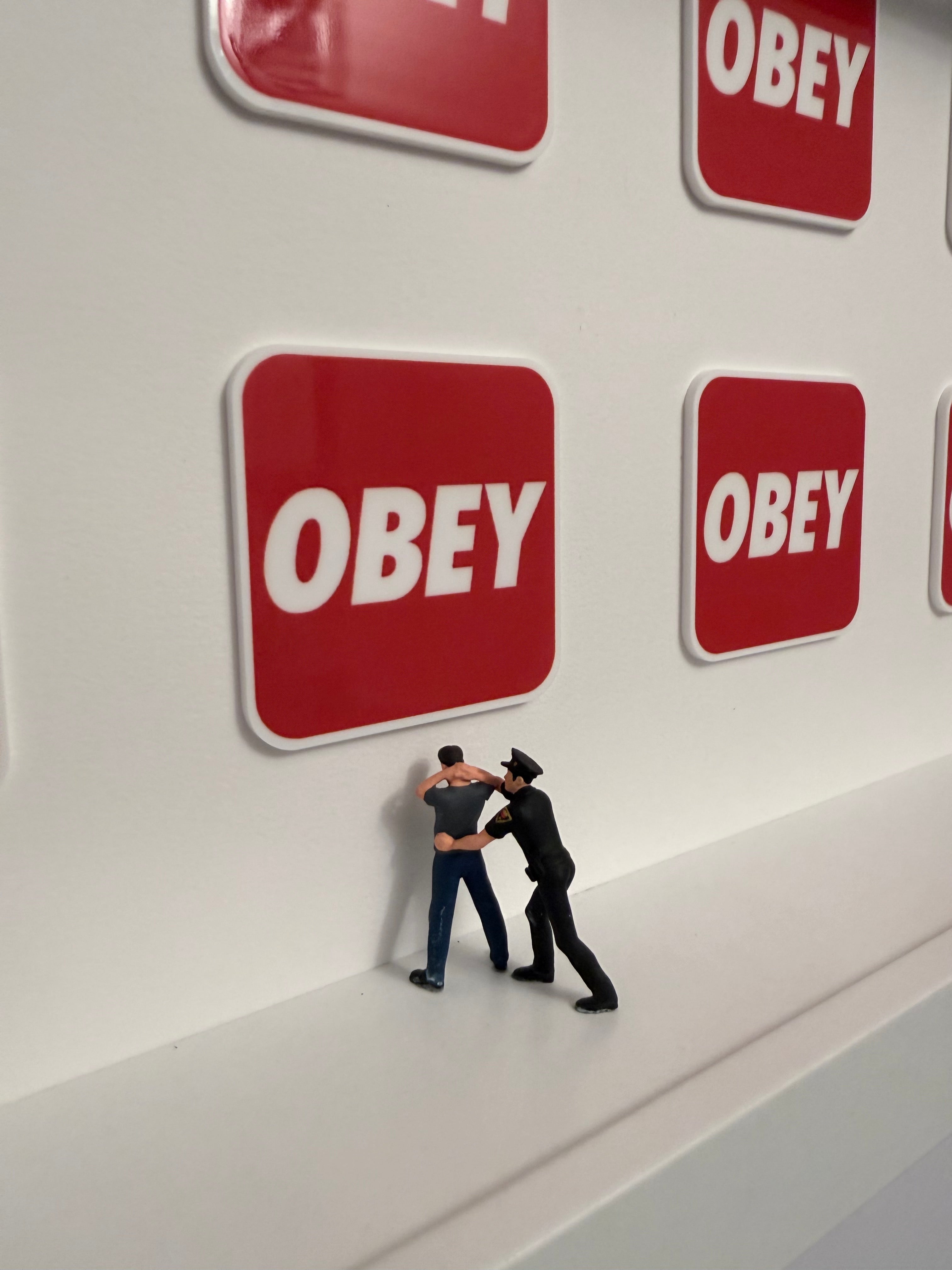 Red 'OBEY' logo on a white surface with two miniature figurines, on being a cop arresting another person.