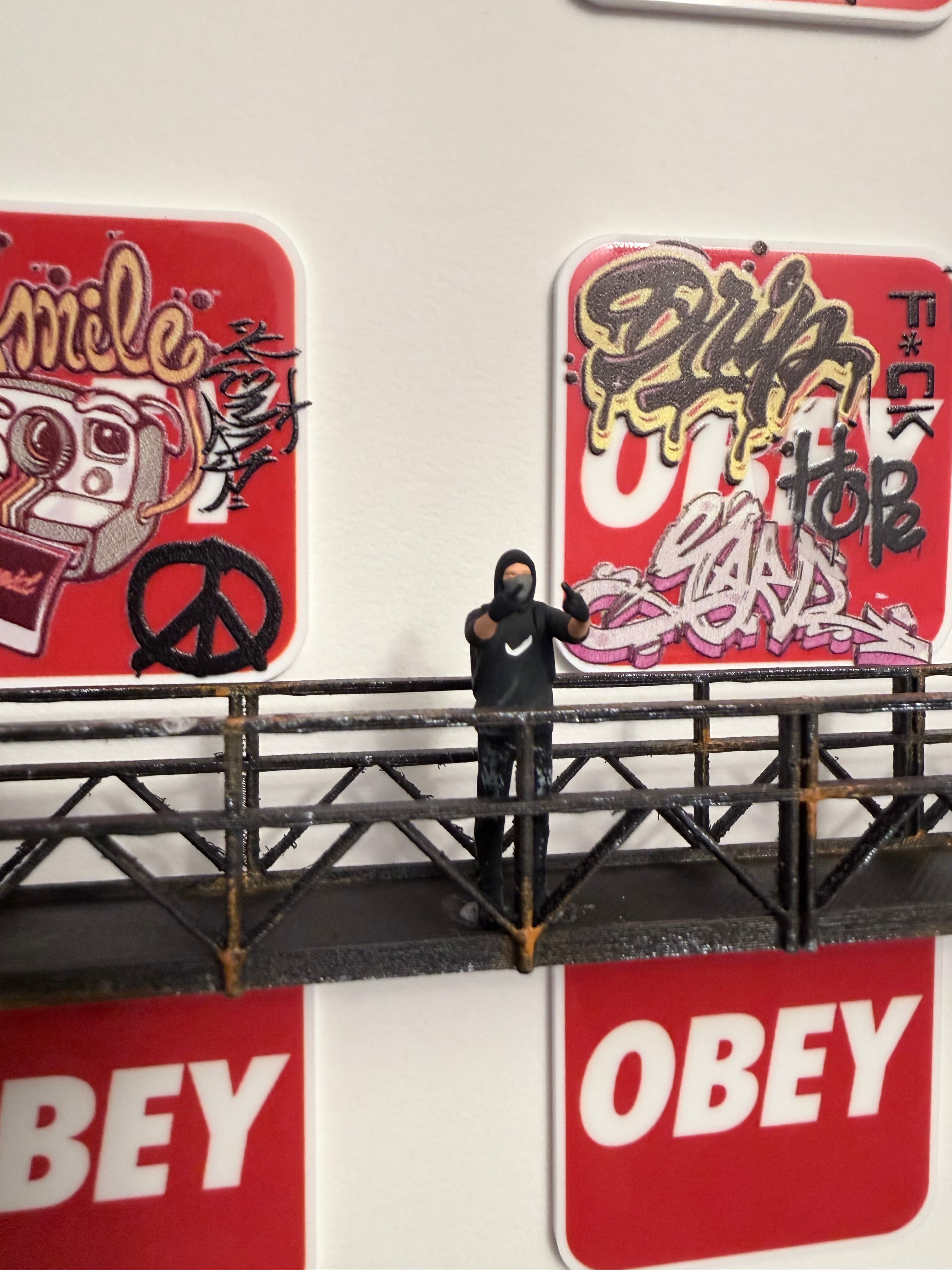 Miniature figurine standing on a small platform showing both his middle fingers, with 'Obey' street art signs in the background