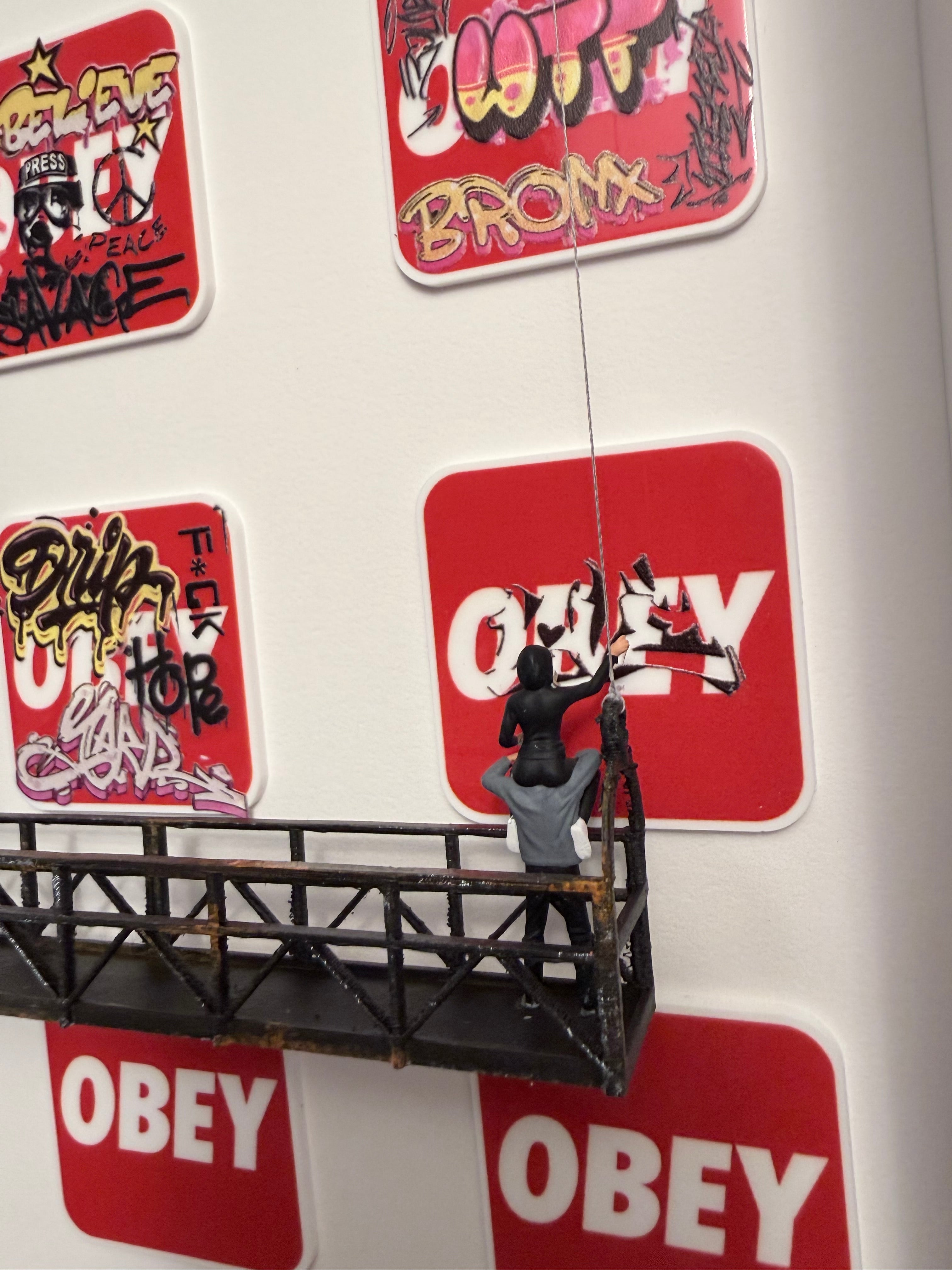 Shadow box, graffiti-covered wall with 'OBey' square logo and two miniature figurines on a platform starting a graffiti.