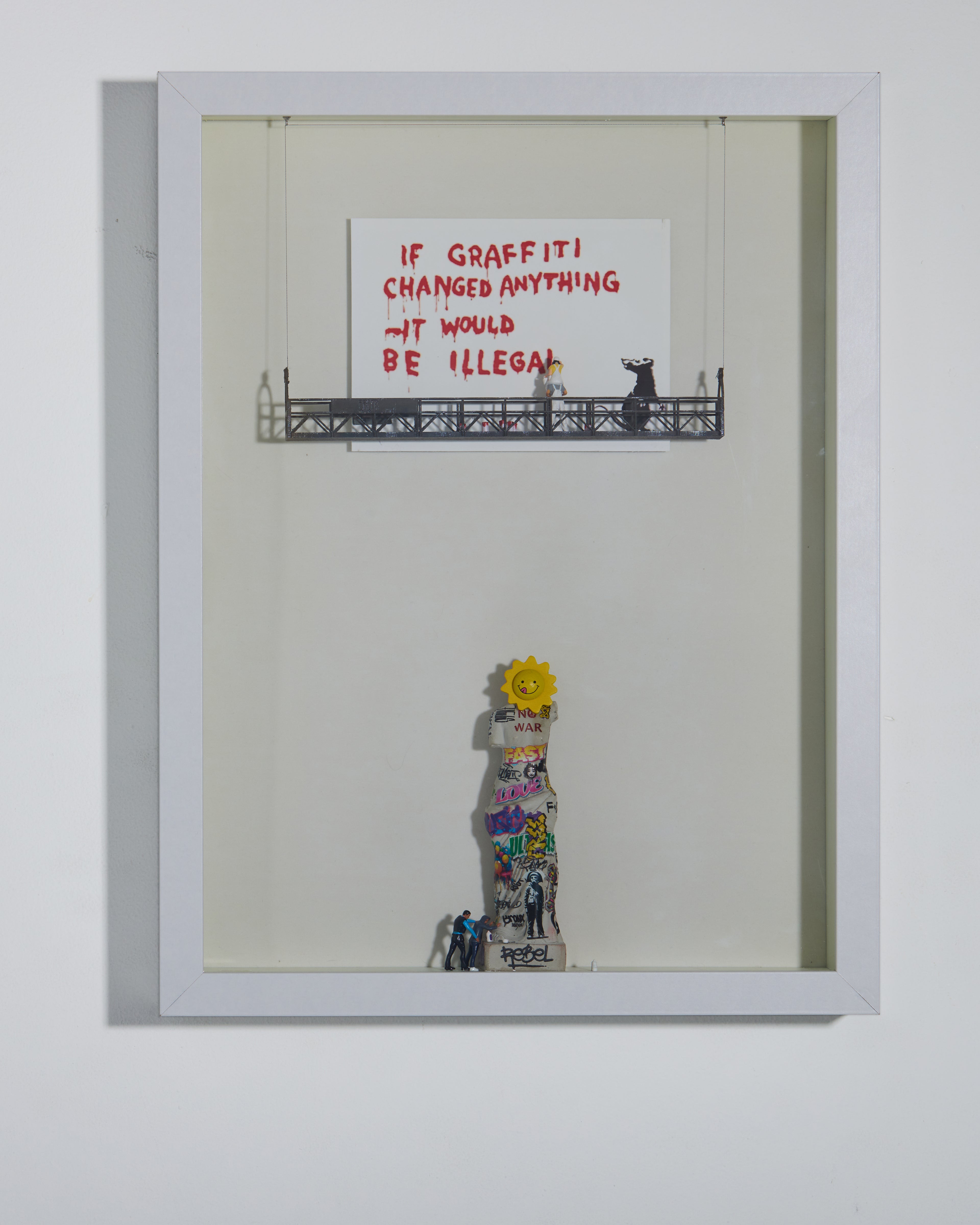 Shadow box artwork with a statue cover by graffiti and text about graffiti legality from Banksy. Miniature figurines are on a platform. 