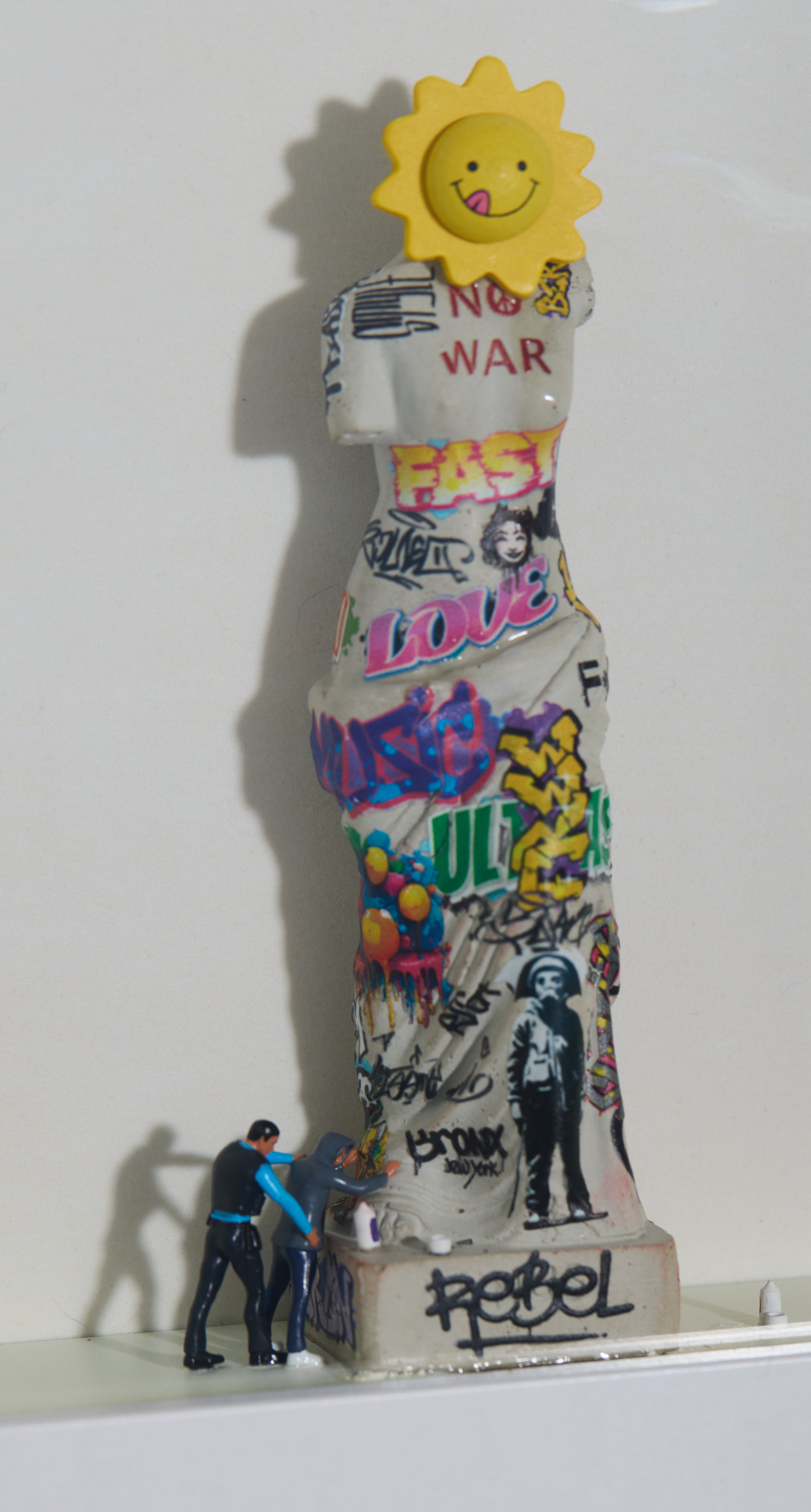 Statue with colorful graffiti and a sun on top, miniature figurines, cop and a criminal are standing next to it.