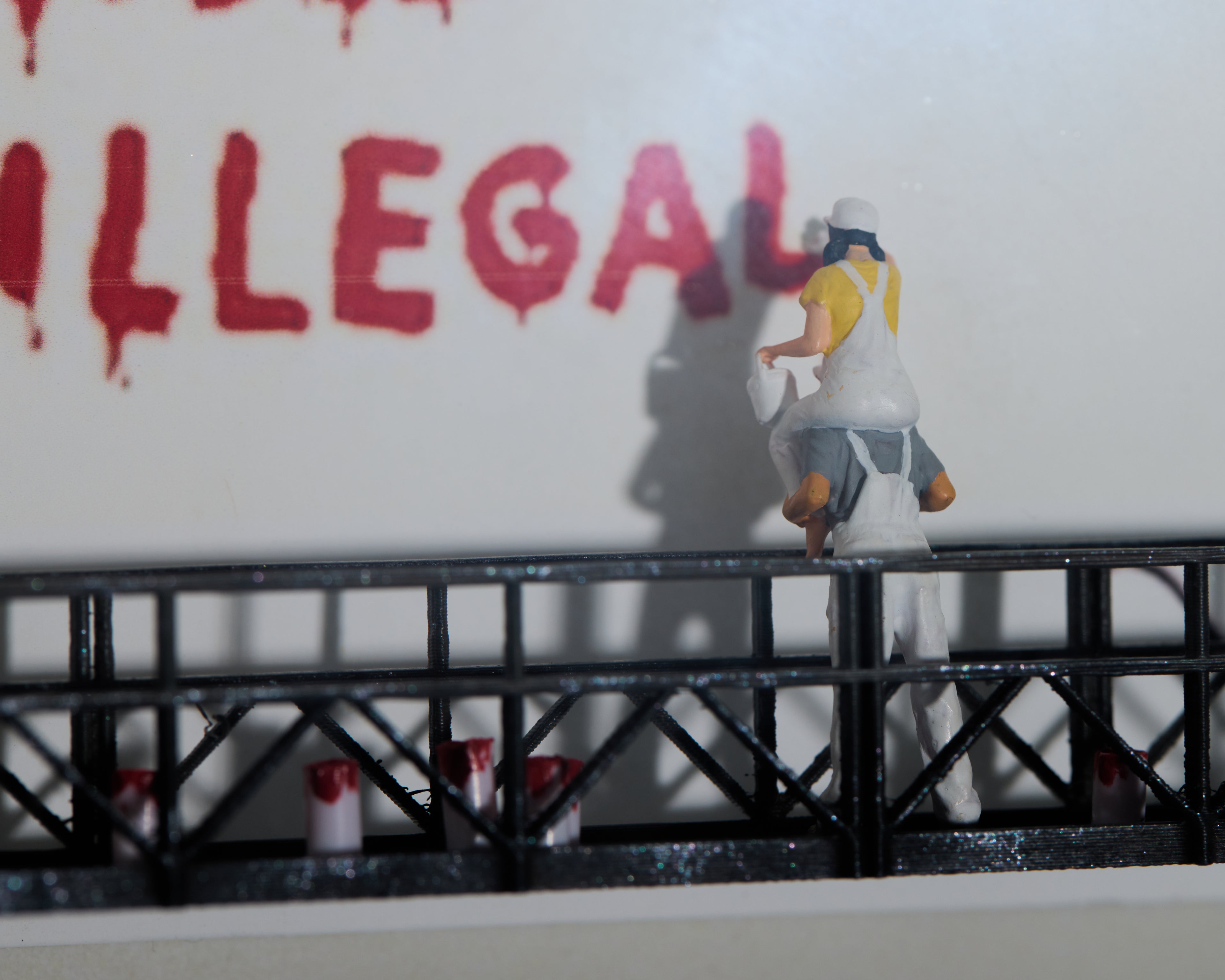 Miniature figurines on a railing with 'ILLEGAL' spray-painted on a wall in red. 
