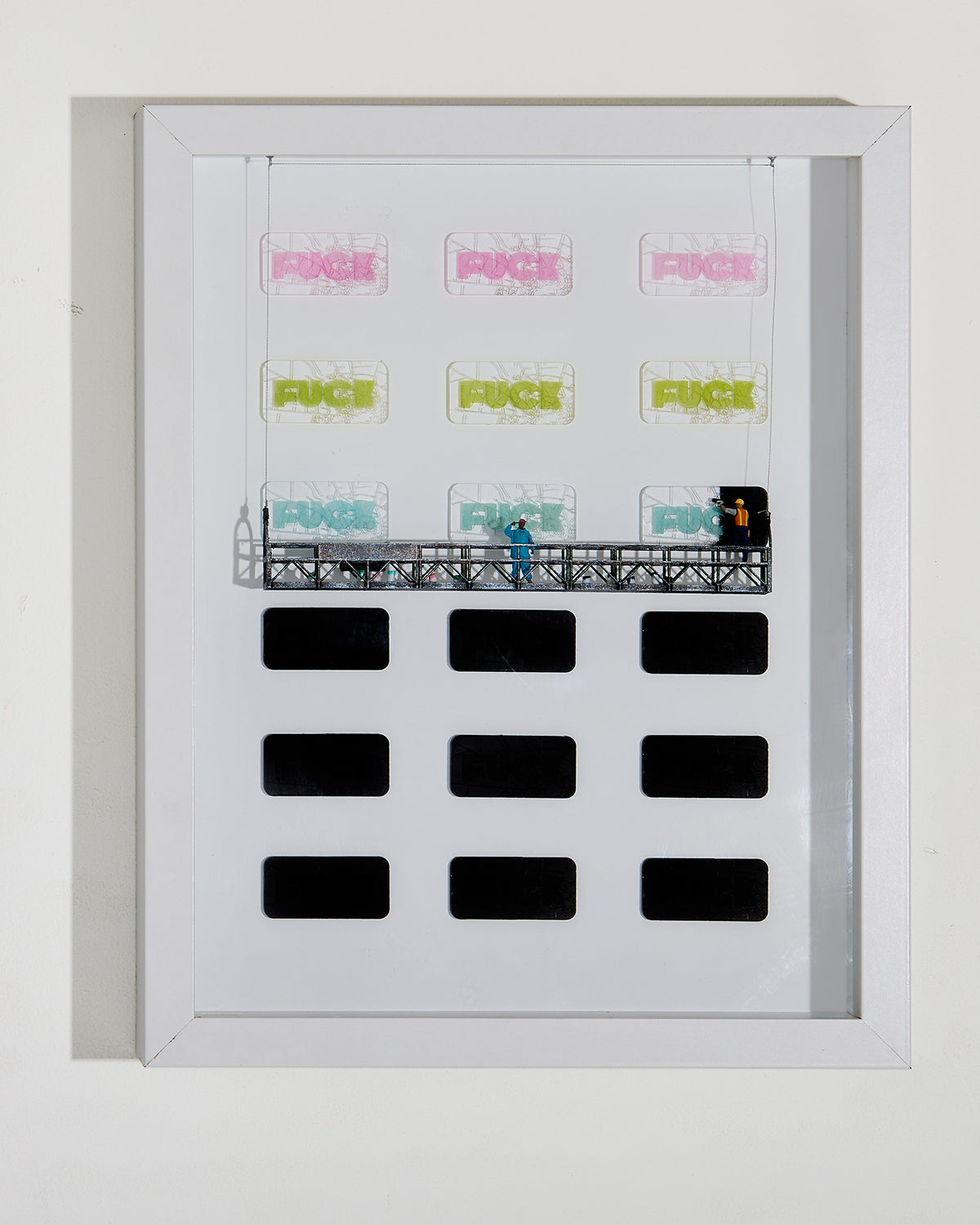 Shadow box artwork, the word fuck is aligned on a white background. A miniature figurine, a painter, is covering all the words in black. Another figurine is on the platform pretending to shoot himself with his hand. 