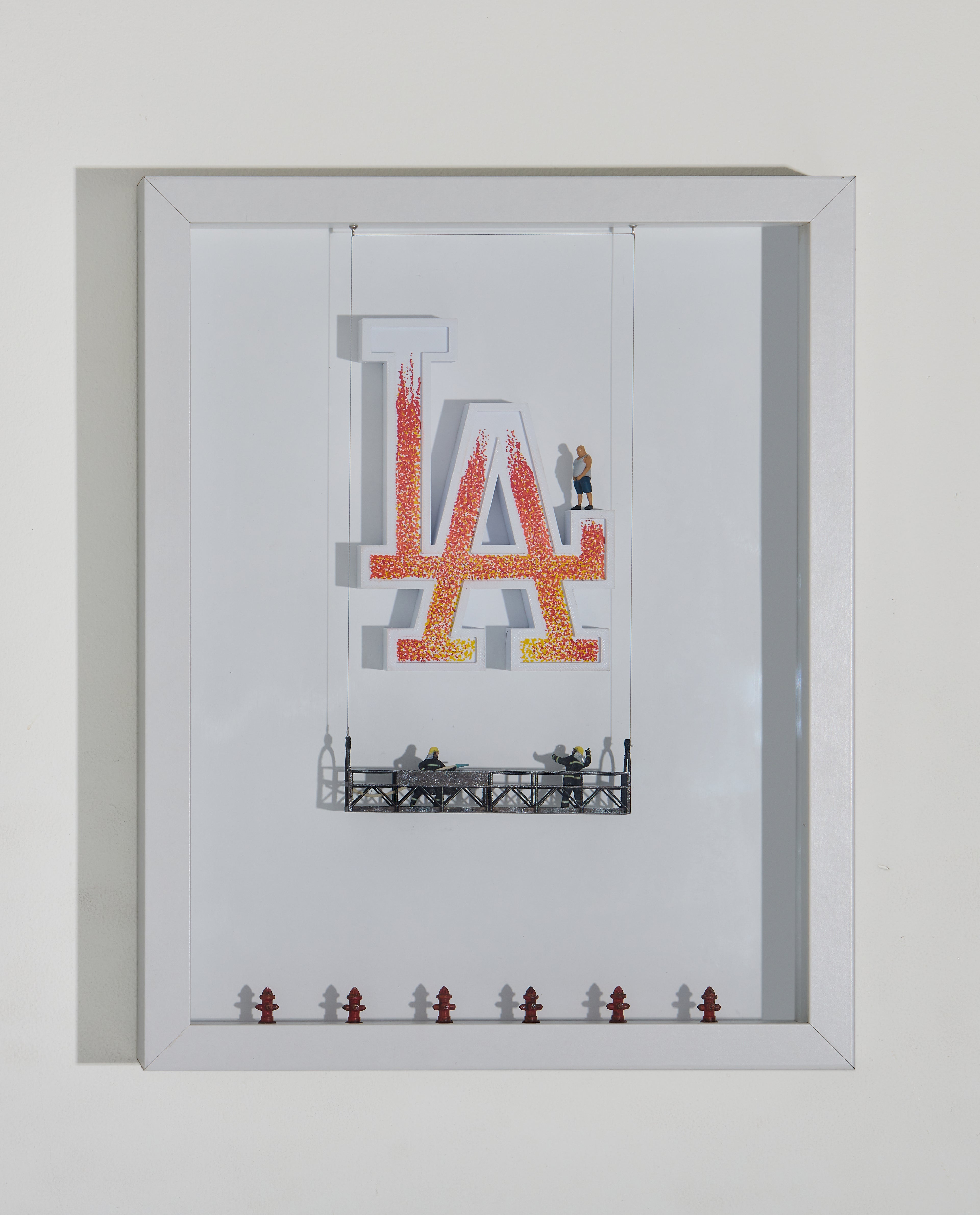 Shadow box artwork with 'LA' sign on fire and miniature figurines. One on the LA logo itself and two miniature firemen on a platform, on a white background