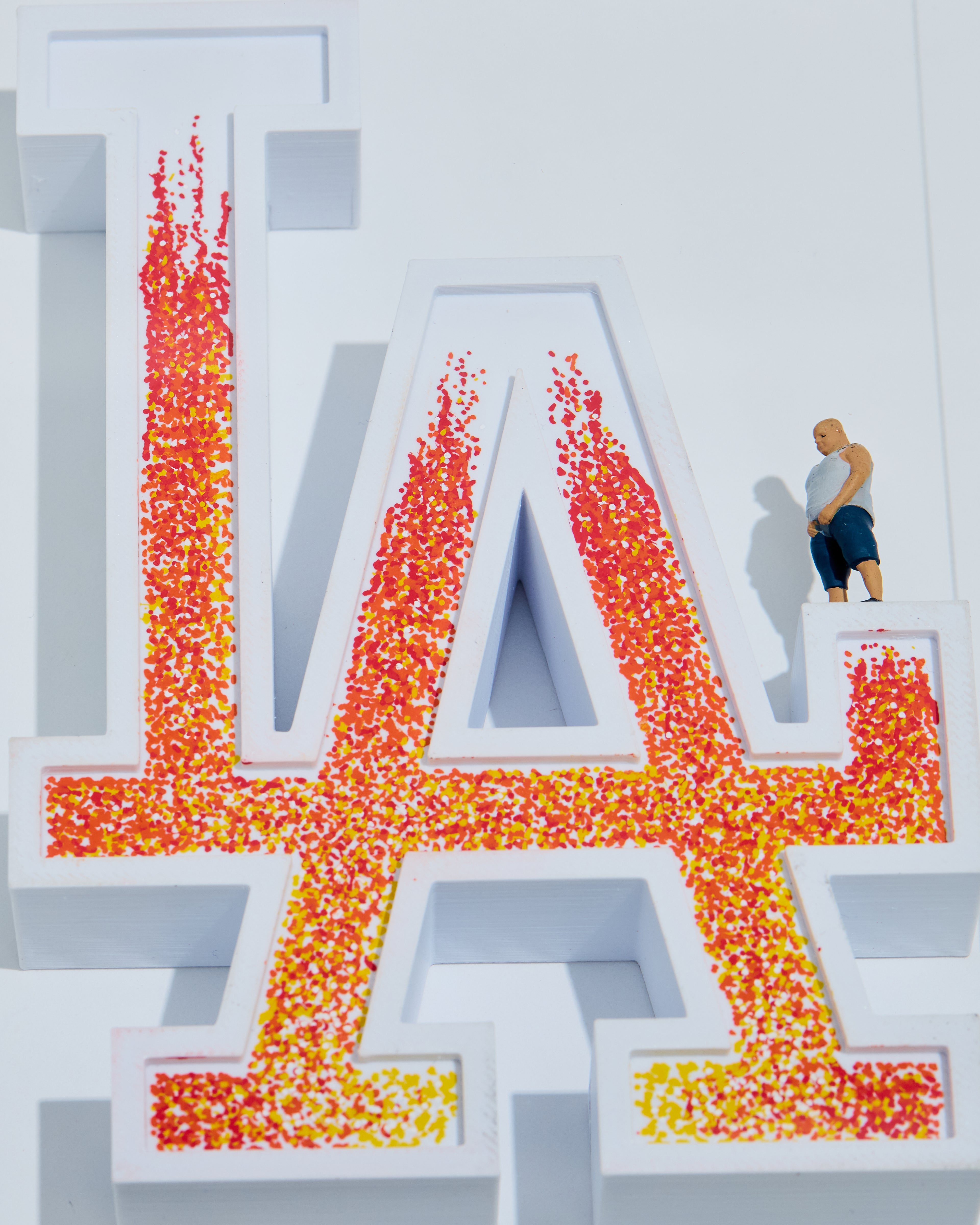 Large 3D letters 'LA' on fire with a miniature figurine standing on the 'A' and peeing to help extinguish the fire