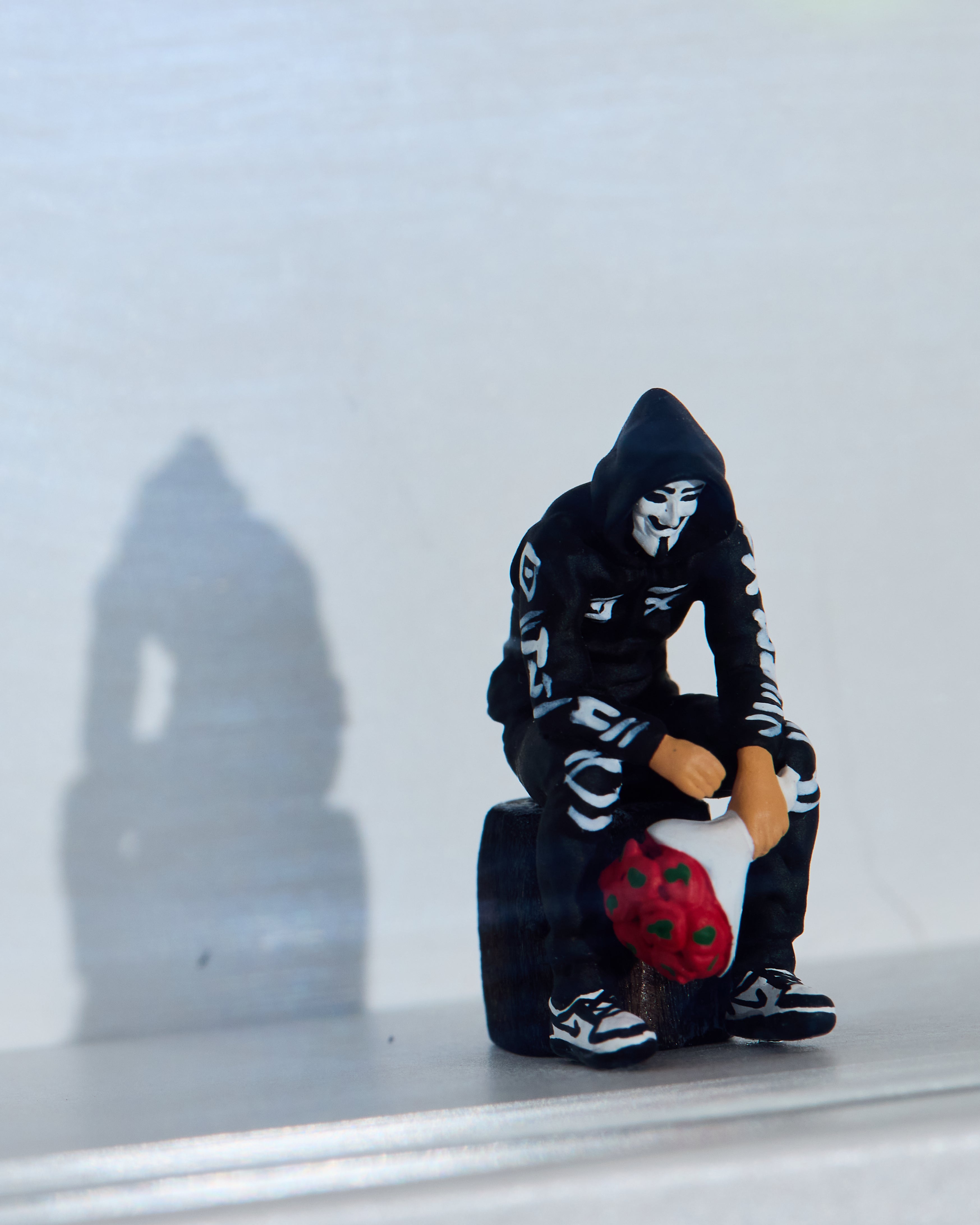 Miniature figurine in a black hoodie with a mask, sitting on a suitcase with red flowers against a white background. In a shadow box.