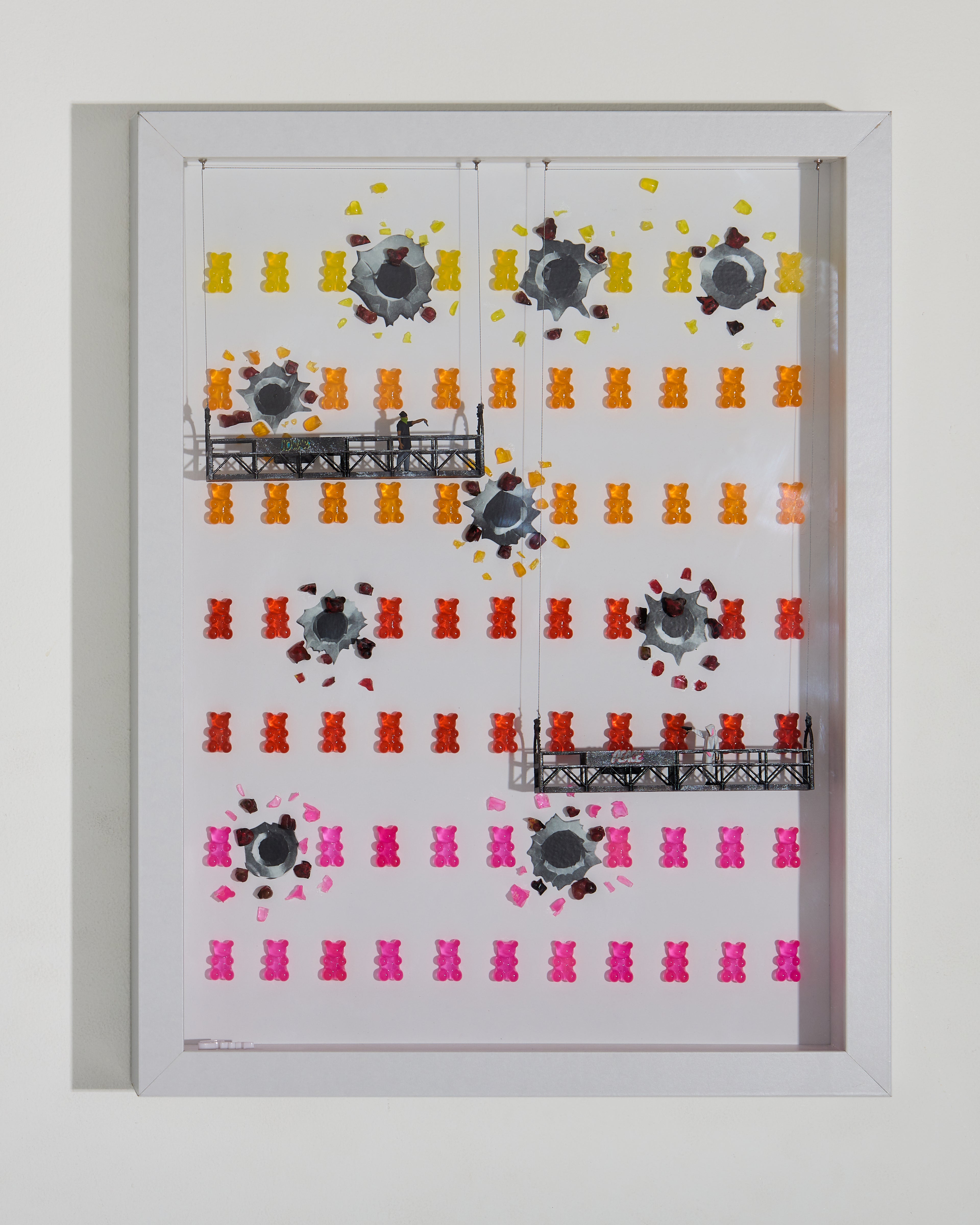 Rows of colorful gummies arranged in lines, some broken with visible bullet impacts. two miniature figurines on lower and upper platforms shoot at each other across the shadow box. 